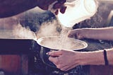 A hand pouring hot water into a pot that another set of hands is holding