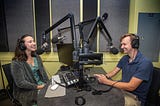 Two people sit in a recording booth talking into microphones.