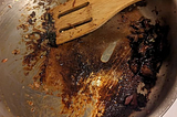 A stainless steel pan with some brown burnt stuff at the bottom and a wooden spatula scraping it up.