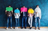 Five adult waiting in line holding various colored speech bubbles made of cut out construction paper convering their faces.