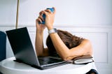 A woman with her head face down on a white table while holding her phone and a her laptop in front of her