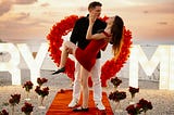 Man holds woman, surrounded by roses and the words “marry me” in lightbulbs.