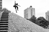 Skater does a flip-trick over a cobbled bank.