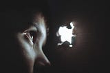 A young person in the dark looks through a small hole in the wall to the light on the outside