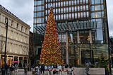 A conical tree with red and gold baubles on it and gold lights all over it in a plaza with an old building to the left and a modern building behind it.