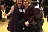Two women next to each other. One is a little person with shoulder length red hair and is wearing a purple sleeveless dress. The other has short brown hair and glasses is wearing a purple jacket and is seated in a wheelchair. Both are smiling.