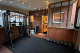 A home gym on a porch, black rubber flooring on most of the ground. A row of kettlebells to the right, a functional trainer with Smith machine on the left.