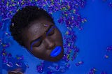 Anthropology in the News. Ancient chewing gum discovery. Image: Black woman in blue water with blue eye and lip makeup.