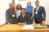 Four Davidson College students dressed in business professional attrie, stand behind New York State Supreme Court Justice J. Machelle Sweeting ’93 who is sitting at a wooden desk. An American flag is hanging next to the wall behind the group of five.