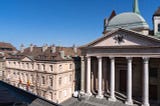 A view of the MIR museum next to Saint-Pierre Cathedral. Classical European architecture with a foreground of a building featuring a neoclassical façade with tall columns and a triangular pediment. In the background, rows of old rooftops with chimneys against a clear blue sky.