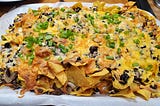 A big pile of tortilla chips, taco meat, sliced mushrooms and cheese topped with green onions on a parchment paper line baking sheet.