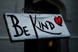 A white rectangular sign on a black post with black letters reading BE KIND with a red heart outlined in black.
