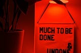 banner reading “much to be done & undone by @rayoandhoney hangs from a sconce with an orange bulb next to a plant.