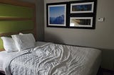 A hotel room bed with three pictures on the wall beside it: one of mountains, one of a lake, and one of a meadow adjoining a forest