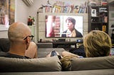 Família sentada no sofá, de costas para a foto, assistindo televisão na sala de estar de seu apartamento.