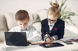Two children dressed professionally using laptops and tablets indoors.