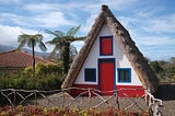 A triangular-shaped thatched house with white wall, red door and green shutters; a rustic wooden fence runs in front, and two palm trees.