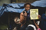 A Black person sits in front of their tent smiling, with their thumb up next to a sign that reads “Mayor Breed we will gladly trade this tent for a hotel room!”