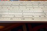 A close-up of a wooden slide rule, showing various logarithmic scales and numerical markings. The slide rule has patent numbers printed along the top edge and includes scales labeled with numbers such as 1.3, 1.35, and 1.4. The background features a colorful surface with handwritten text and designs.