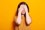 A stressed female covering her face and eyes with her palms. She is wearing a yellow jumper against a yellow background