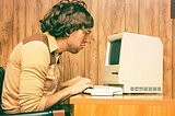 Image of a man with scruffy hair hunched over a vintage computer in a room with wood paneling. The man’s face is scrunched.