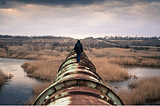 A human figure walks along a rusty pipeline in a bleak, drab, wetland.