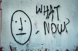 Black graffiti on a white wall — A simple, straight face with the words ‘what now?’