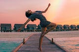 A person practising a stretched pose, on a beach pier with a rainbow in the background.