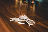 Wood grain bar, with empty 1/4 pint glass, empty of liquid but containing a few ice cubes, sitting atop a stack of two one dollar bills and a paper receipt.