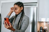 Photograph of a young Black woman with long dreadlocks wearing a gray turtleneck. She stands in her kitchen, holding her phone in one hand, the other is on her forehead. Her eyes are closed. She looks upset.