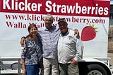A woman and two men are standing in front of a billboard on the side of a trailer with a red and white banner saying Klicker Strawberries. There is a painted red strawberry on the right side of the photo.