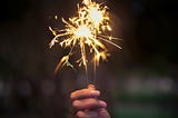 A hand holds two lit sparklers on a dark, blurry background