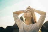 Girl with long brown hair and glasses wearing a white tee shirt. She is outside, with her face up to the sun, eyes closed, smiling. Hands are on top of her head.