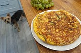 My dog Luna has come to the kitchen attracted by the smell of the freshly cooked omelette. The omelette appears on the right to the picture on a white plate. Some green grapes can be seen at the top of the picture, on the same worktop as the omelette.