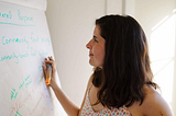 A white woman with shoulder-length brown hair wearing a flower-print camisole holds an orange marker and gazes at a white flip chart with indeterminate green writing.