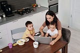 A mom sitting at the kitchen table working from home on her laptop with her 2 kids around her.