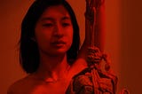 A mostly red photograph of an East Asian woman with straight, dark hair. She is bare faced and is looking at a rock being suspended with rope. She is wearing a necklace with a pendant that reads Felicity.