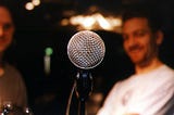 A close-up of a microphone, with two blurry listeners in the background.