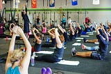 A large group of people sit on the floor of a cavernous gym. They are reaching overhead in a stretch, with an instructor standing up and leading them.