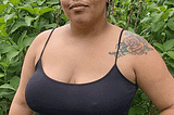 Isabela é uma mulher negra, e está com o cabelo crespo castanho preso em um coque. Ela tem uma tatuagem de flor no ombro esquerdo. Está usando brincos de argola, uma regata preta e uma saia amarela estampada. Isabela está em pé com plantas ao fundo.