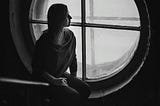 Grayscale photo of a woman sitting near a round wooden framed window
