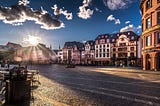 A photo of a plaza with cobblestone pavement and old-looking buildings with southern German architecture. The sun is low on the horizon with clearly distinct rays beaming across the square, creating long shadows on the pavement.