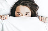 A scared woman lying on bed covered with a white blanket with only her eyes and forehead exposed