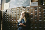 A woman in a blue dress leaning back against an archived stack of documents, looking a little sad.