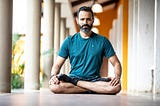 A man sits cross-legged in a meditative posture on the floor of a bright corridor, wearing a teal athletic shirt and black socks. He appears calm and focused, with natural light highlighting the peaceful setting. Reflecting the perspective of Ramneek Bhogal on integrating wellness into busy professional lives.