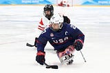 Josh Pauls is a white man wearing a white helmet with a metal face cage and a navy blue hockey jersey with USA and a letter C indicating that he is the team captain on the wrong. The number 27 is written on the shoulder. He is sitting in a sled hockey sled with a carbon fiber casing cpvering his lower body decorated to look like an American flag. He is a double above the knee amputee. He is holding two sticks, and using one to control the puck. A player from Canada is in the background.