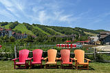 Five Muskoka chairs (2 red, 3 orange) lined up against the backdrop of a ski hill and blue skies in the summer.