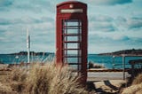A red telephone booth standing empty in front of the shore.