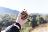 A person’s arm is outstretched and is holding a compass in front of a scenic view of sand, mountains, and shrubbery in the backgound.