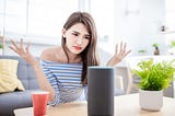 A woman sitting in a living room looks at her smart speaker with disdain and raises her hands in frustration.